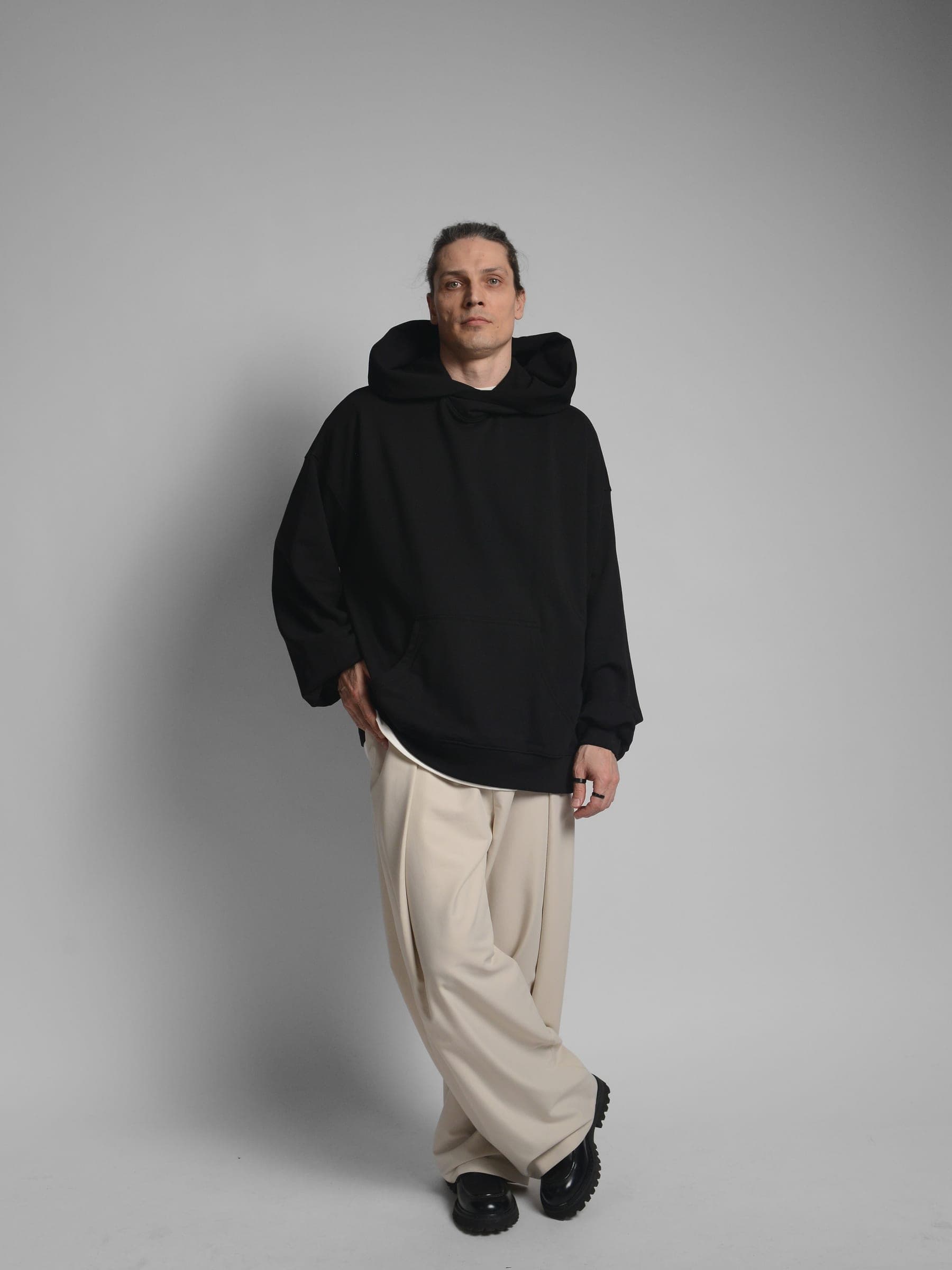 365.APParel Oversize Hoodie Black — man, full body side view, holding large tote bag, hood visible, cream wide-leg trousers, studio