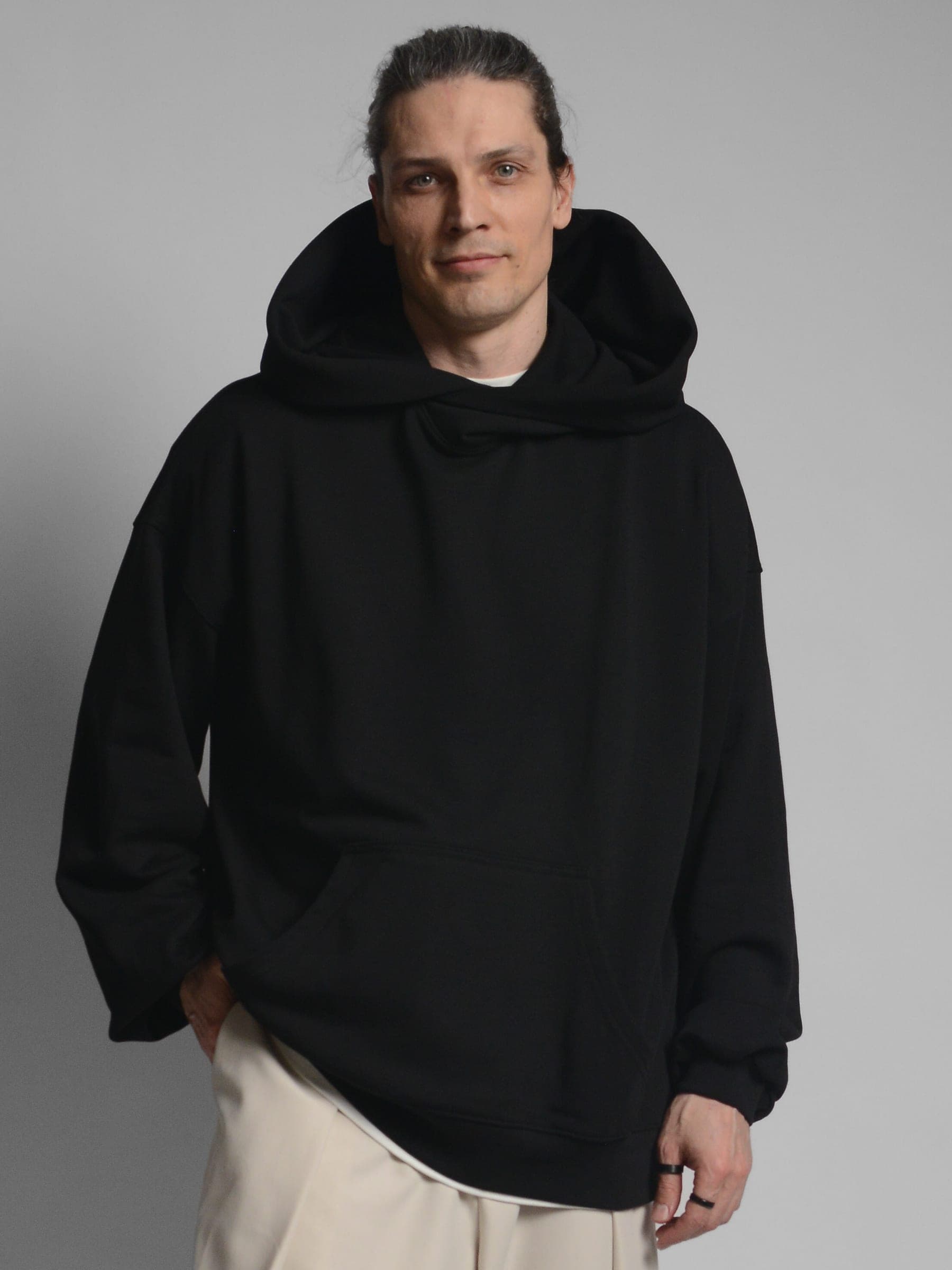 365.APParel Oversize Hoodie Black — man, full body, three-quarter view, holding large tote bag, hood visible, cream wide-leg trousers, studio