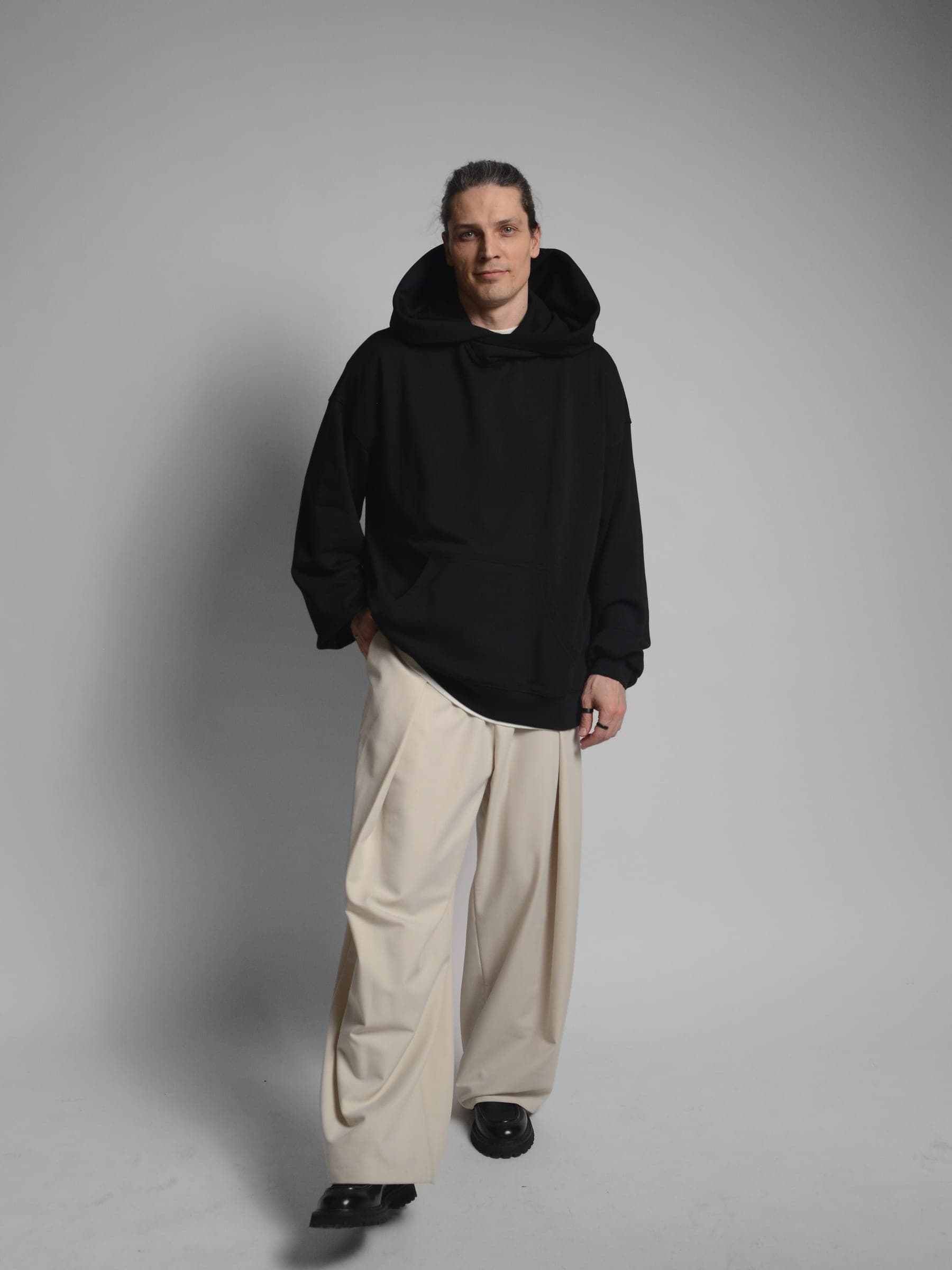 365.APParel Oversize Hoodie Black — man, cropped back view, hood visible, holding tote bag, one hand on hip, studio