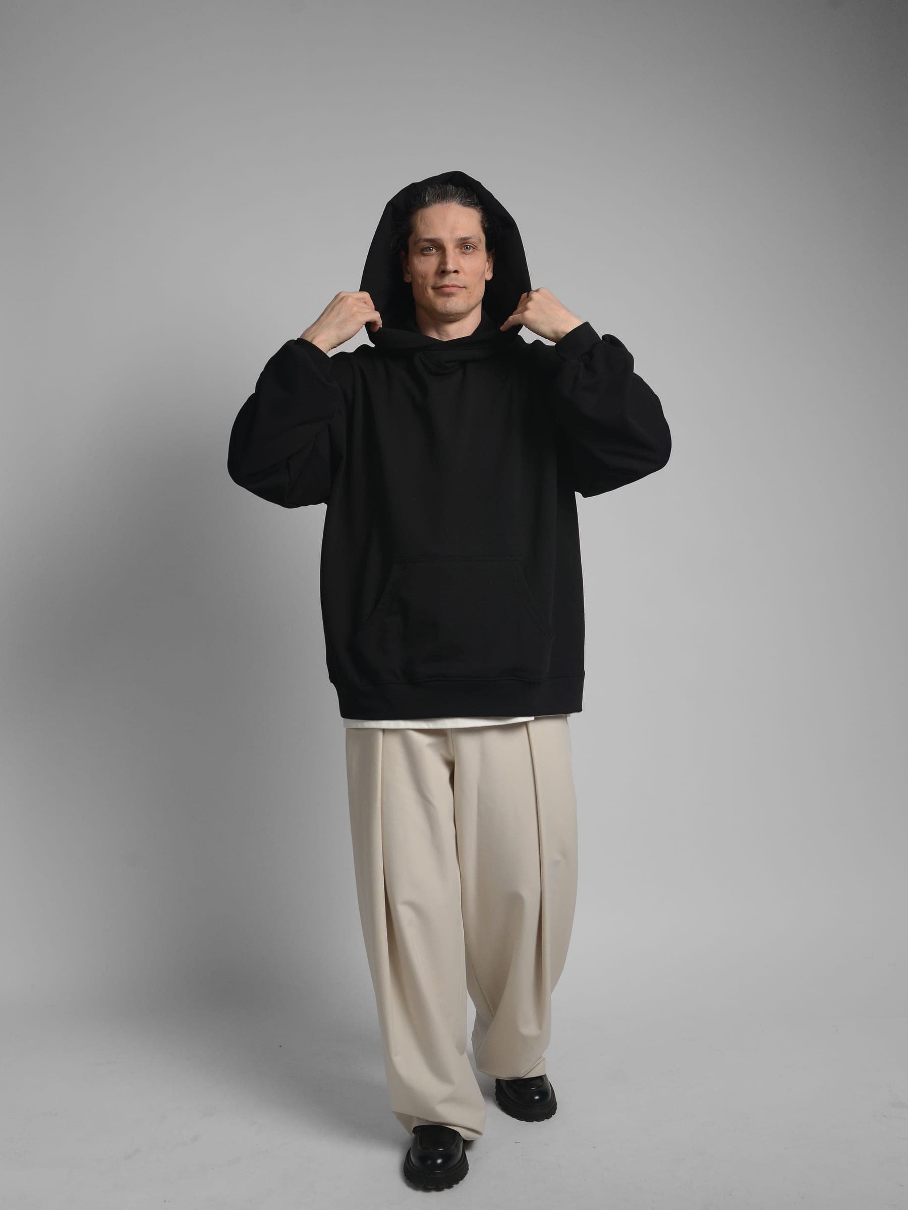 365.APParel Oversize Hoodie Black — man, full body side view, arms raised showing sleeve, hood visible, cream wide-leg trousers, studio
