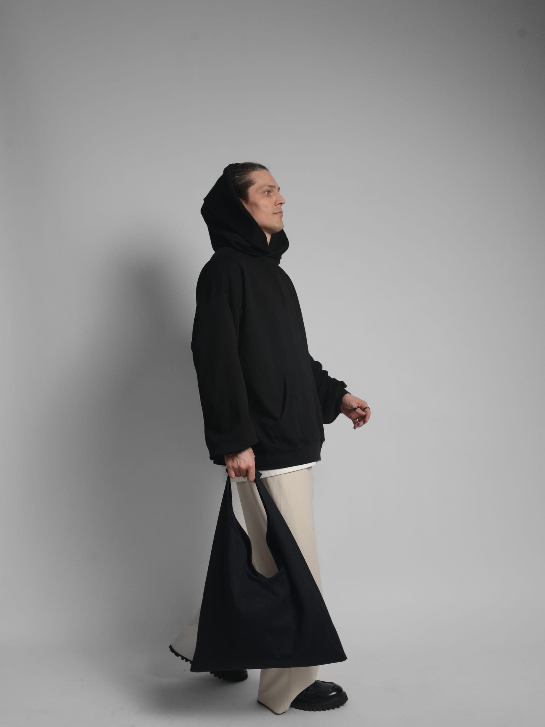 365.APParel Oversize Hoodie Black — man, full body front, smiling, hands in kangaroo pocket, hood visible, cream wide-leg trousers, studio