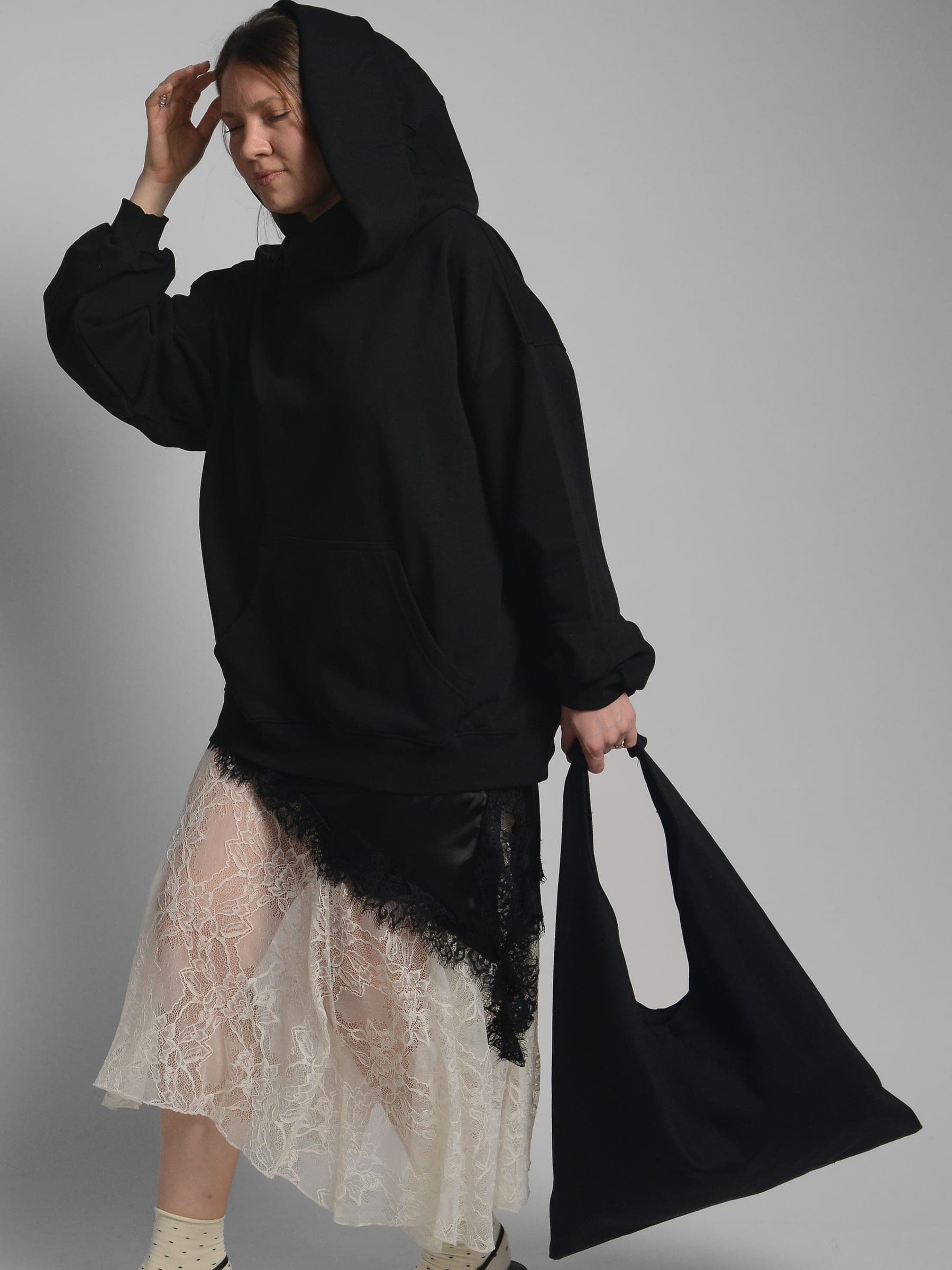 365.APParel Oversize Hoodie Black — woman, full body, hood up, holding large tote bag, lace skirt, looking down, studio