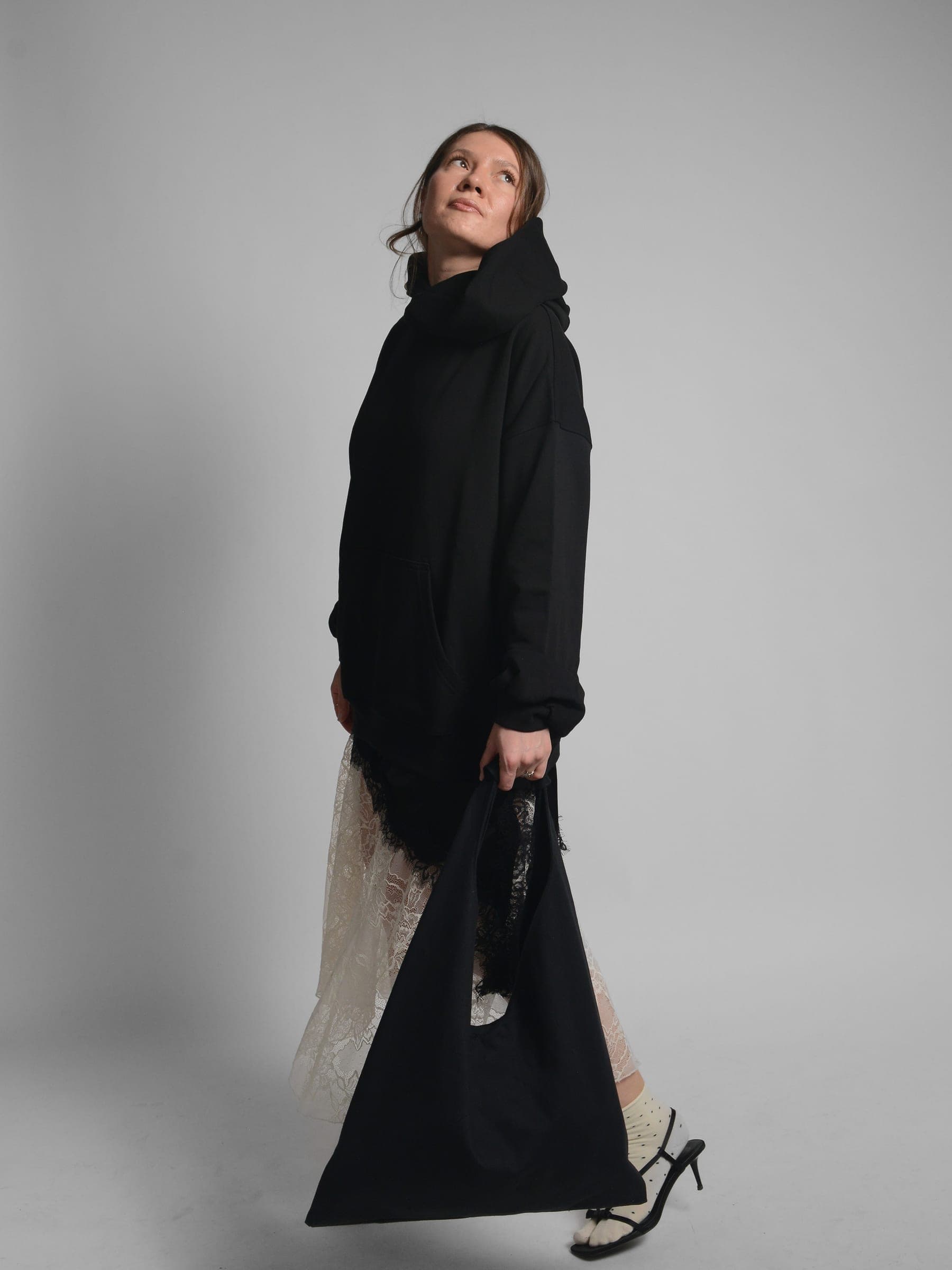 365.APParel Oversize Hoodie Black — woman, full body side view, looking up, hood visible, holding tote bag, lace skirt, studio
