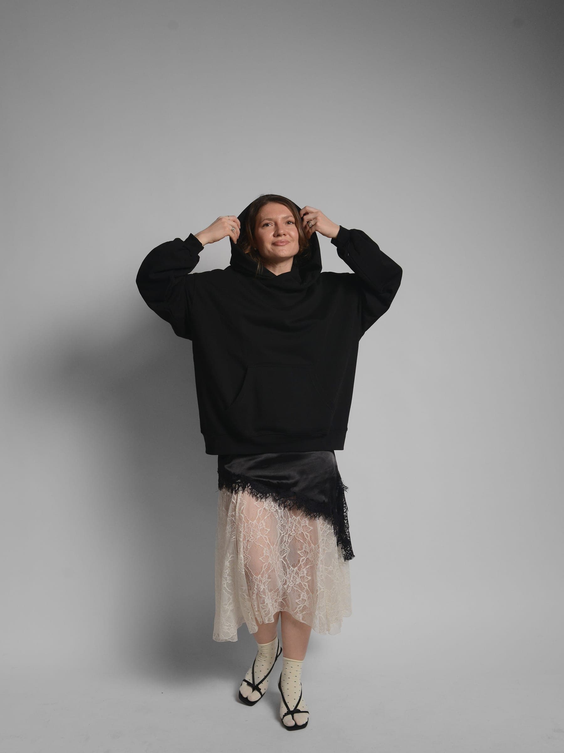 365.APParel Oversize Hoodie Black — woman, full body front, smiling, pulling up hood with both hands, lace midi skirt, studio