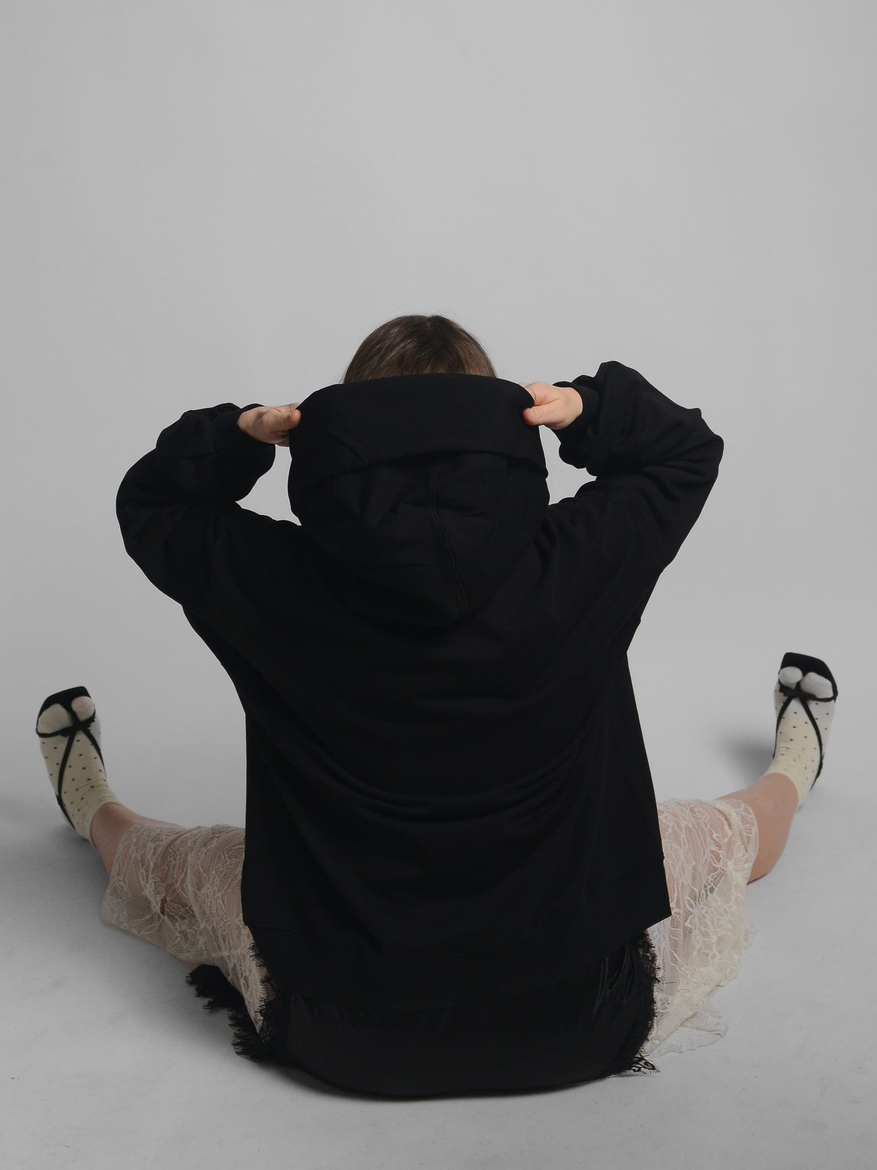 365.APParel Oversize Hoodie Black — woman, sitting on floor, back view, pulling up hood, legs spread, lace skirt, studio