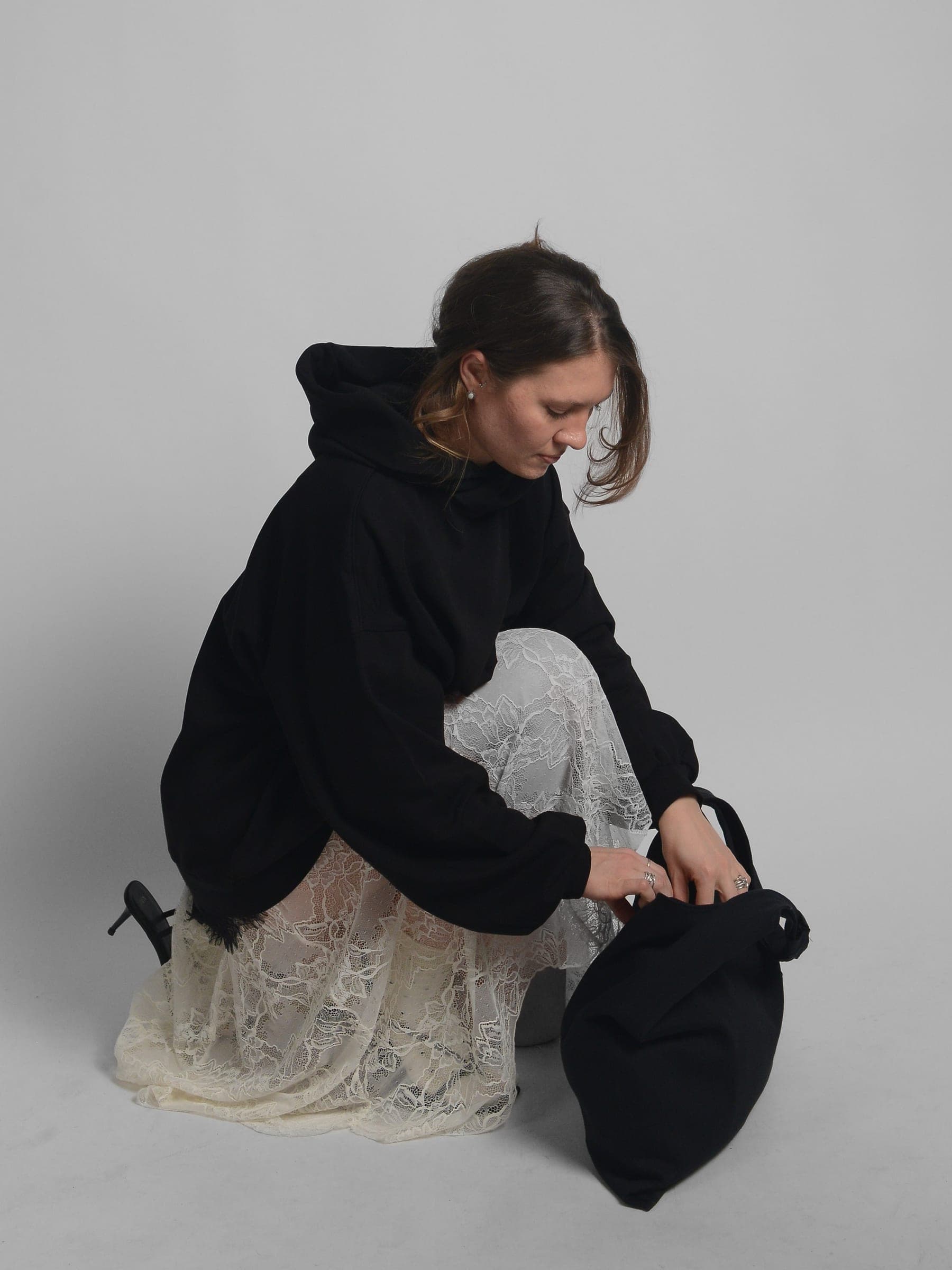 365.APParel Oversize Hoodie Black — woman, crouching on floor, holding black knot bag, lace skirt, hood visible, looking down, studio