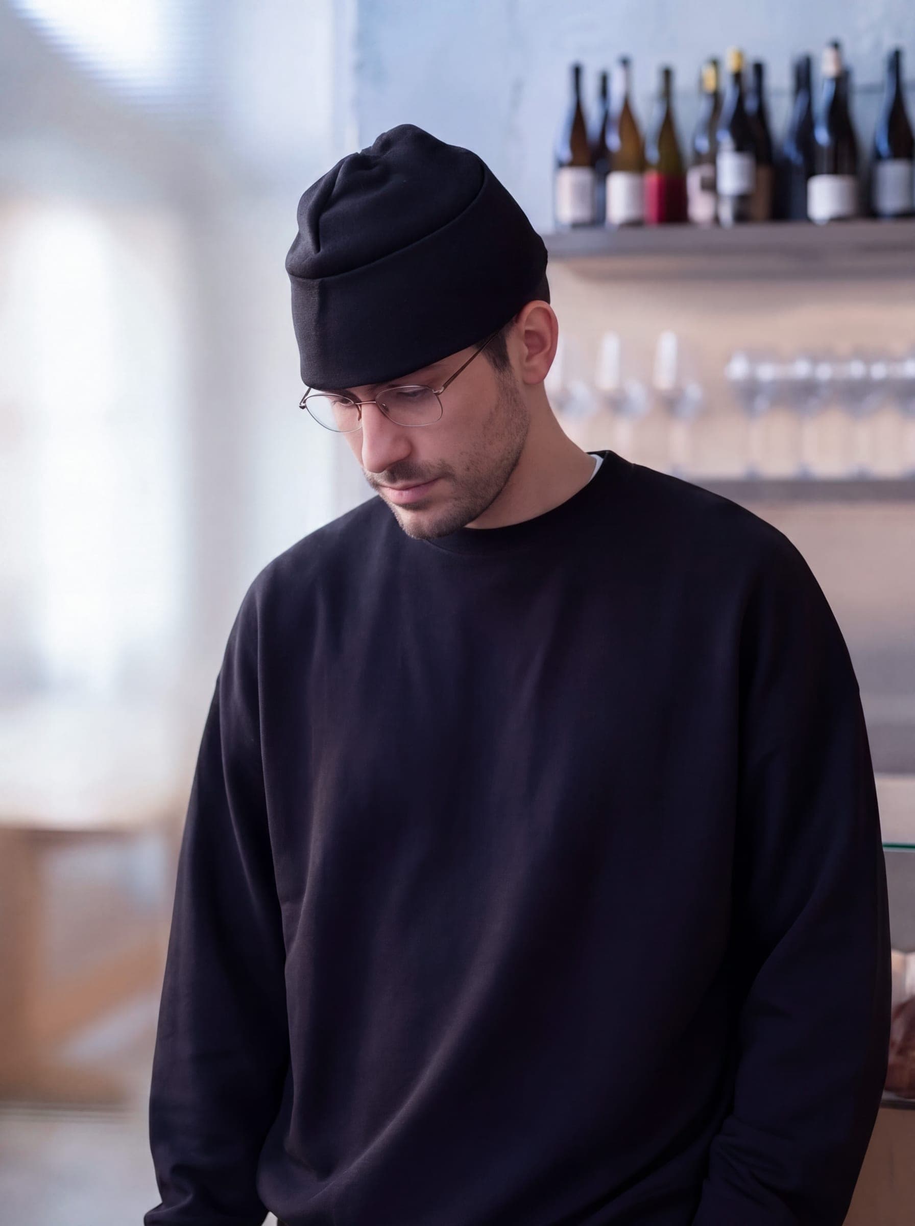365.APParel Oversize Sweatshirt Black — man, cropped front view, looking down, glasses, beanie, wine bar, indoor