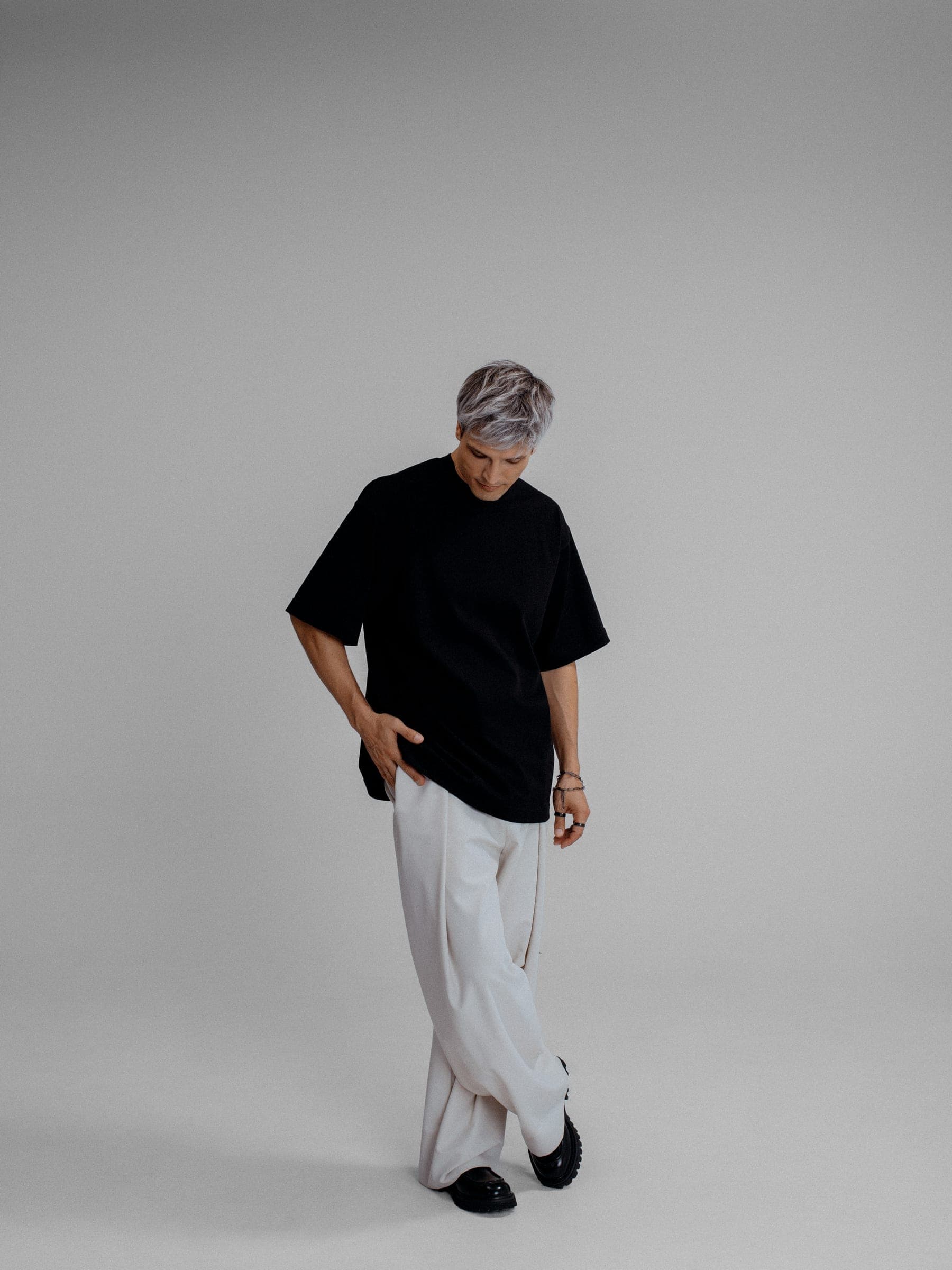 365.APParel Oversize T-shirt Black — man, full body front, gray hair, looking down, white trousers, studio