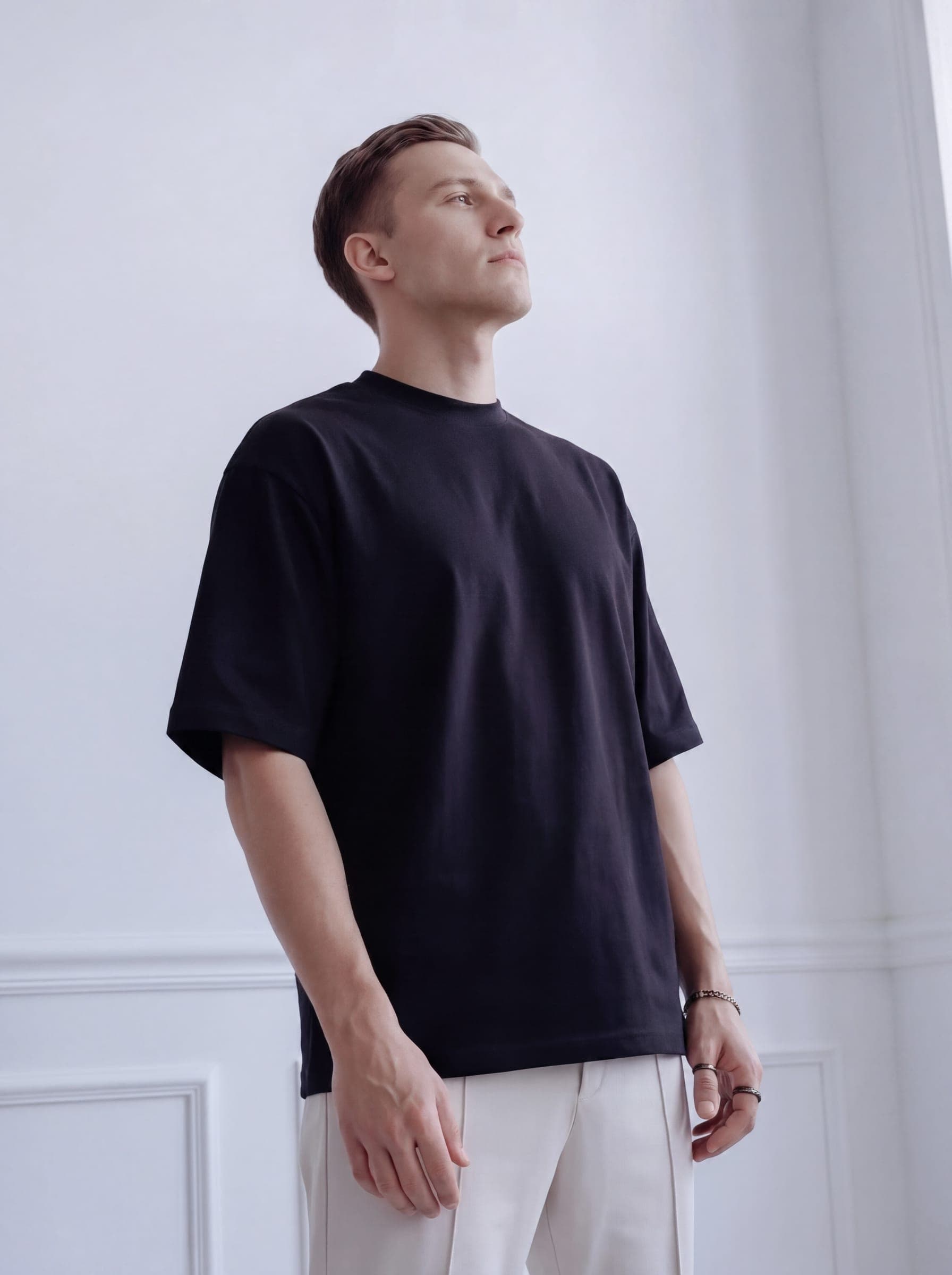 365.APParel Oversize T-shirt Black — man, standing by chair, looking down, full body, indoor editorial