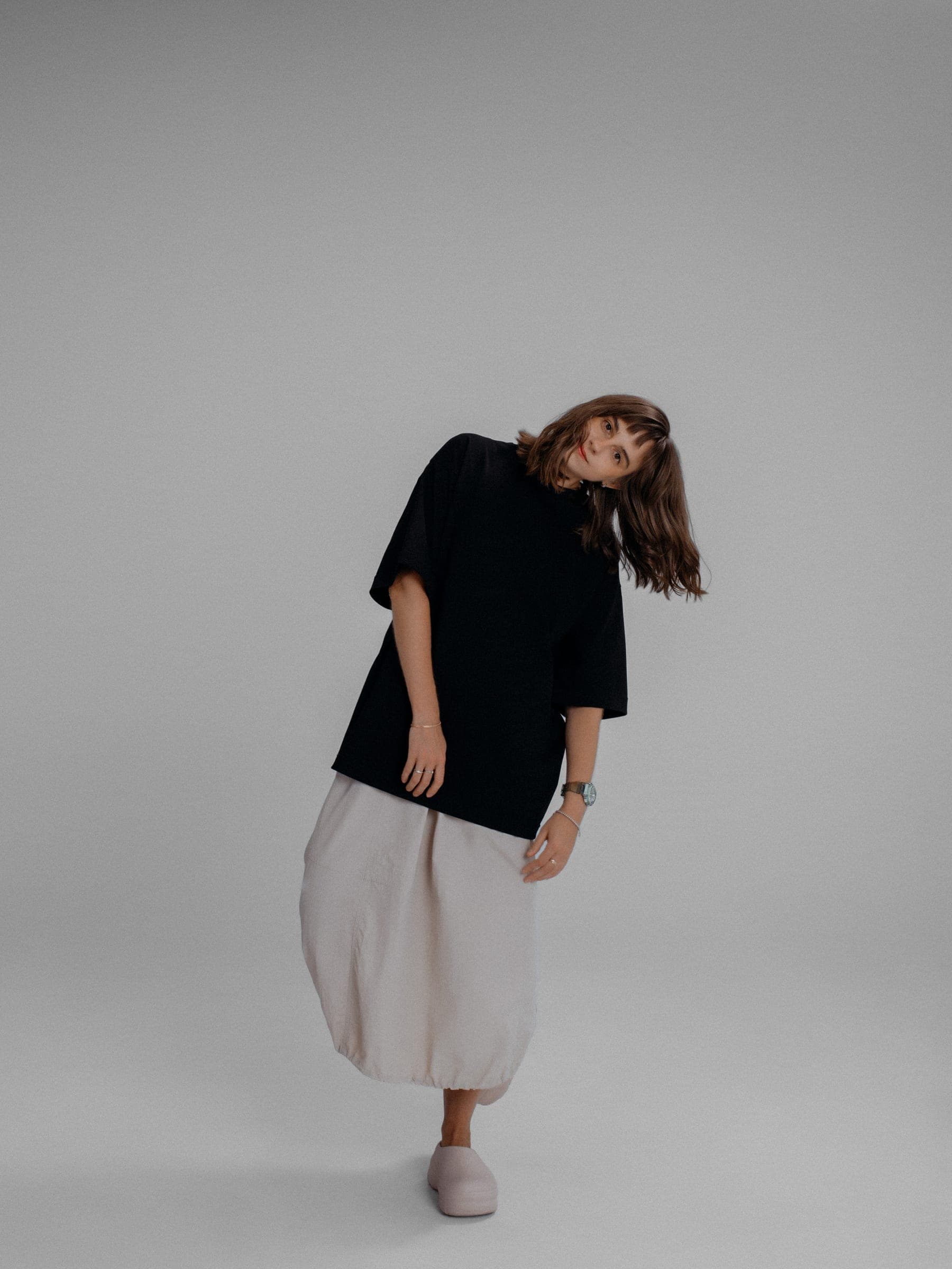 365.APParel Oversize T-shirt Black — woman, full body front, tilting sideways, hair falling, beige bubble skirt, studio